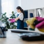 Selective focus of cleaner spraying plant near colleague cleaning telephone on office table